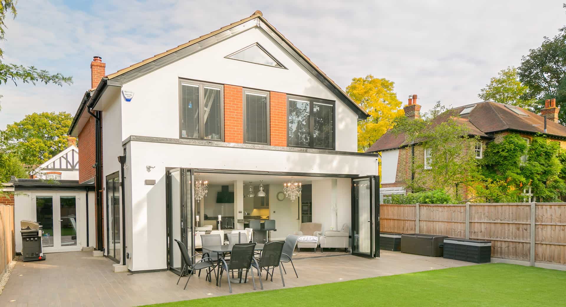 Modern two-story house with large windows and bifold doors opening to a dining area. The exterior features white walls with red brick accents, a spacious patio with dining furniture, and a lawn in the foreground.