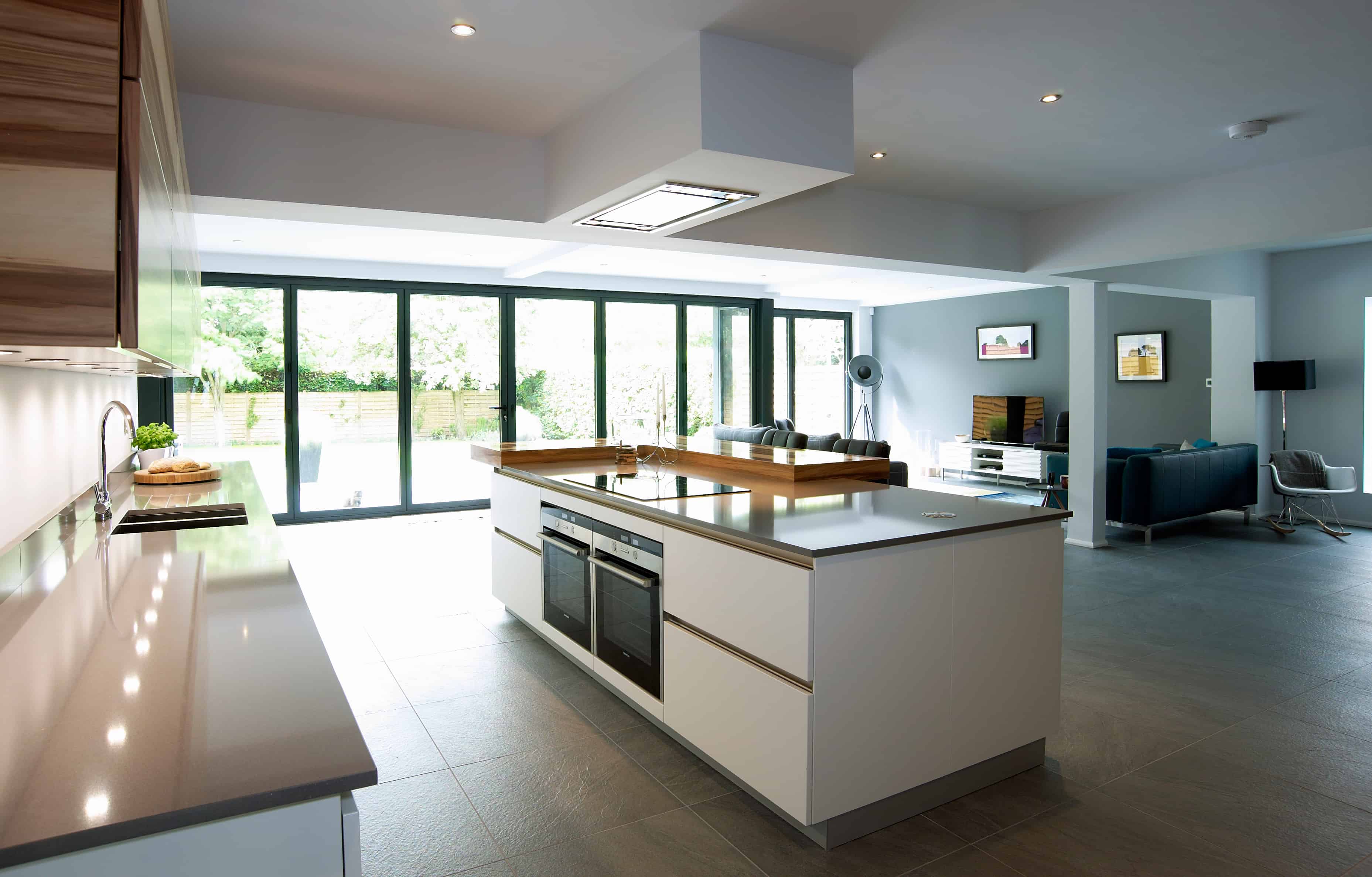 Modern kitchen with a large white island, built-in ovens, and wood accents. The space opens to a living area with large glass doors, letting in natural light. Minimalist decor with neutral colors and sleek surfaces.