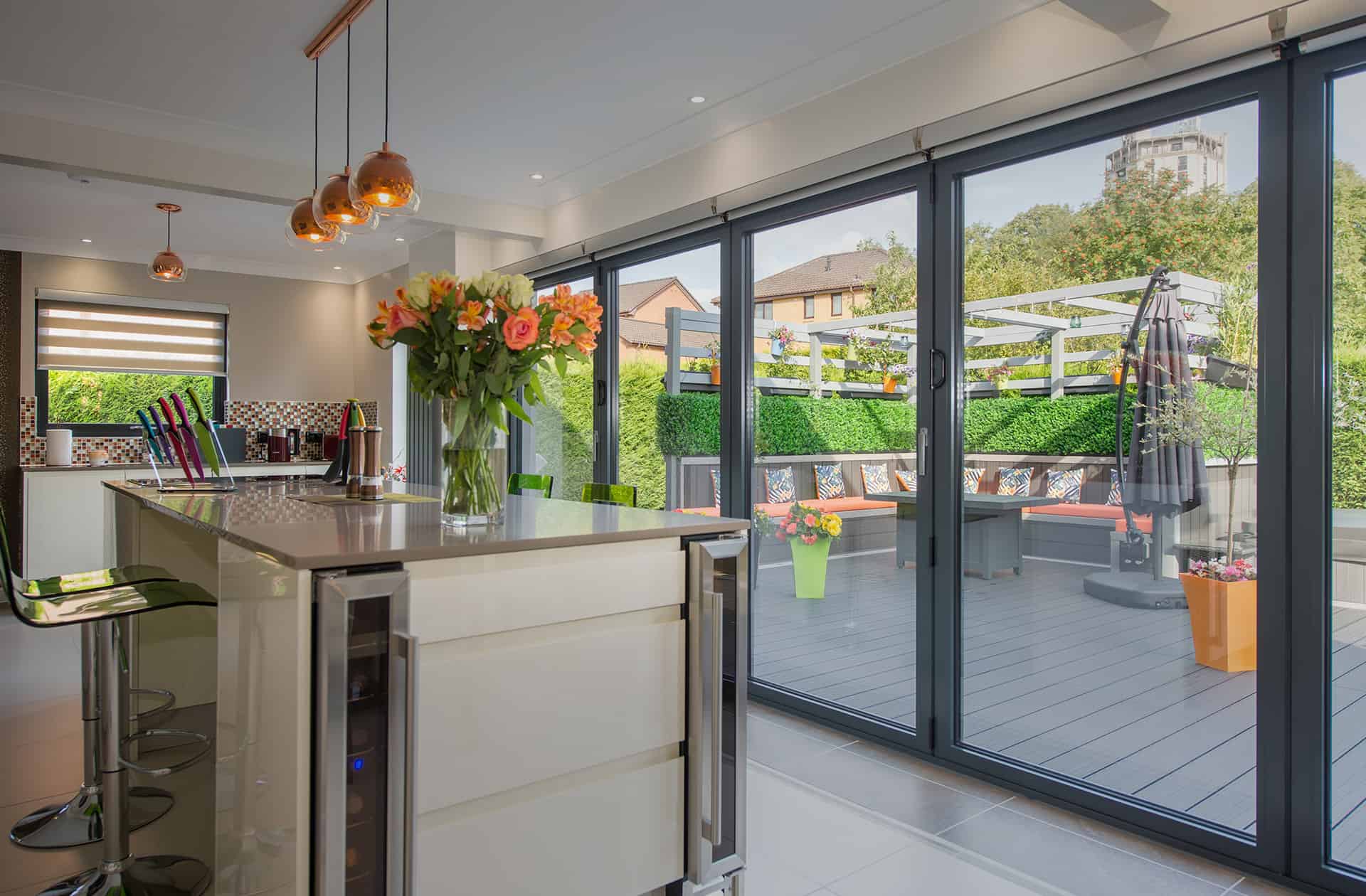 Modern kitchen with an island, bar stools, and a vase of flowers. Sliding doors open to a stylish outdoor patio with seating, greenery, and colorful decor. Natural light fills the space.