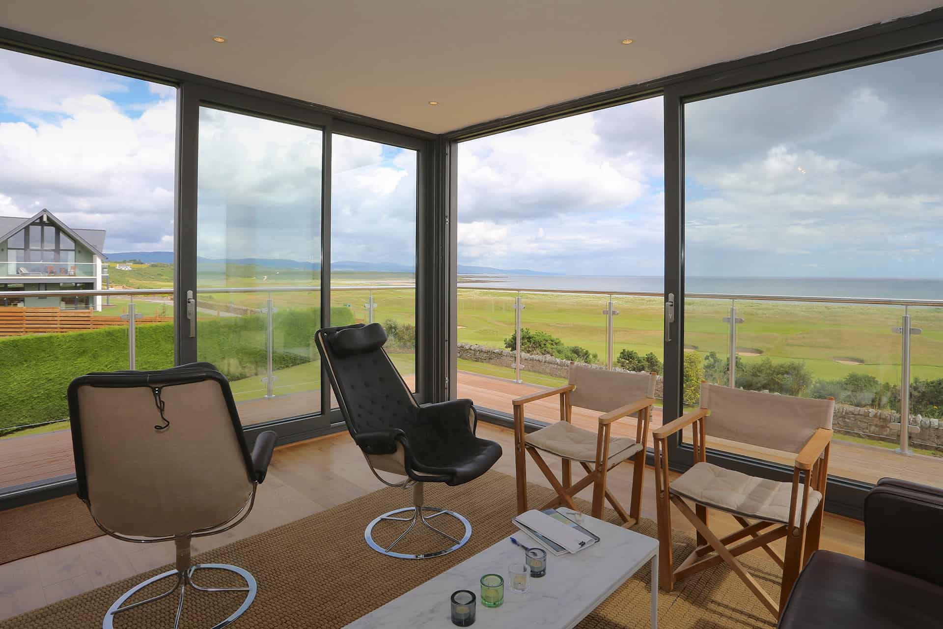 Modern lounge with glass walls and a glass roof overlooking a scenic coastal landscape, featuring two swivel chairs, two wooden chairs, a white coffee table, and expansive views of the ocean and green fields.