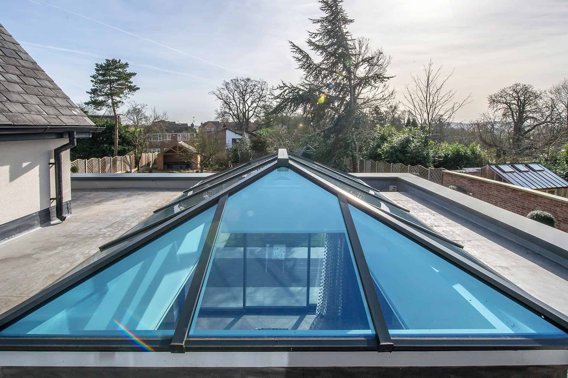 A modern glass roof skylight with a pyramid shape sits on a flat rooftop, reflecting the sky and surrounding trees—an example of bespoke glass solutions—with suburban houses and a wooden fence in the background.