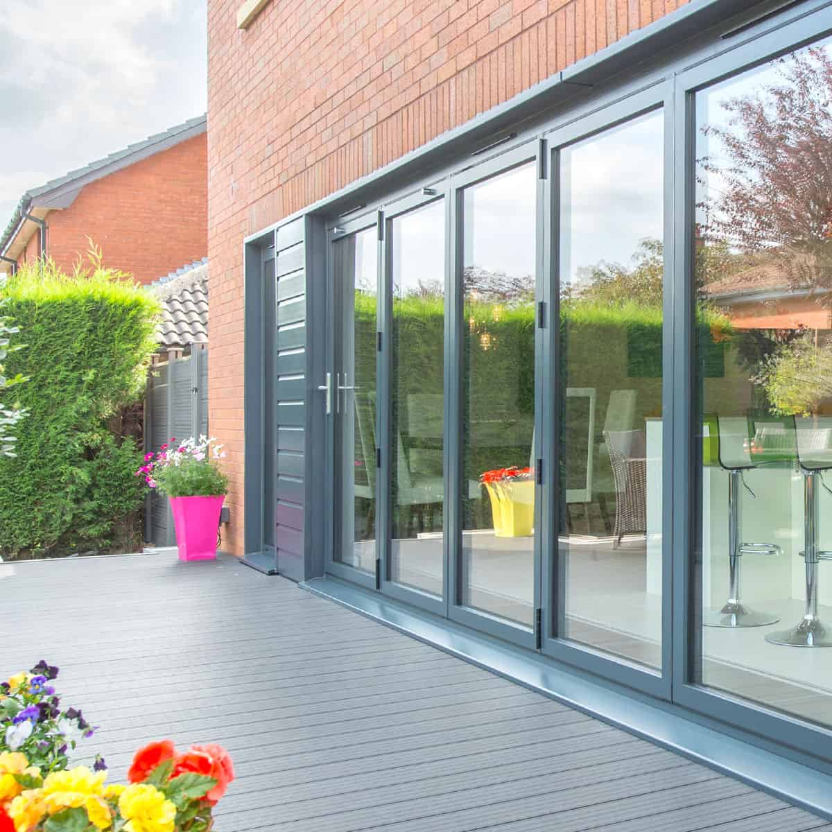 Modern bifold doors open onto a gray patio with colorful potted flowers. The house has a brick exterior, and neatly trimmed hedges are visible in the background.