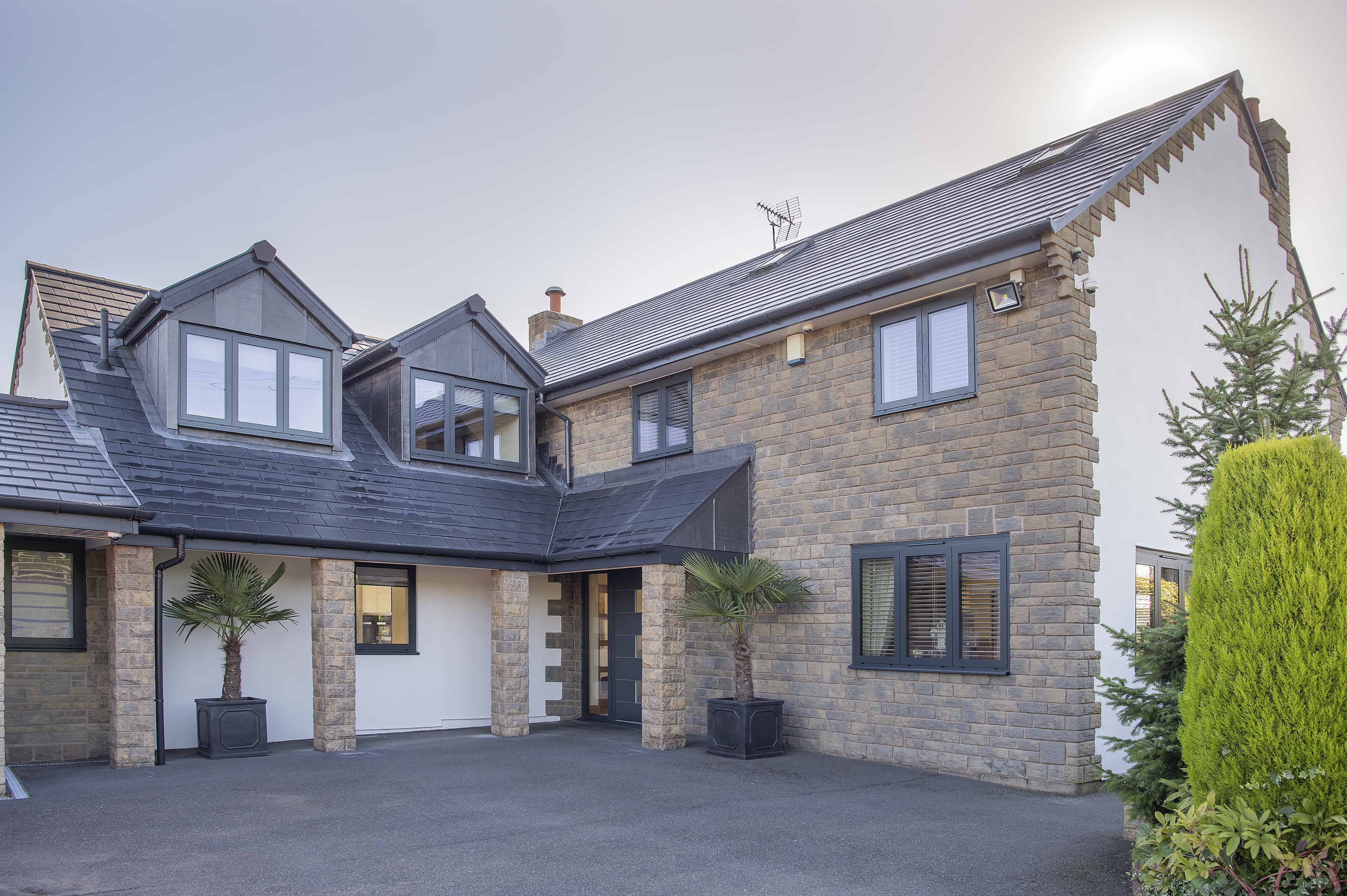 Modern two-story house with stone and white exterior, dark roof, multiple windows, a small porch with two potted palm trees, and a driveway in front. A sleek bifold door opens to the outside, while the sun shines above the roof.