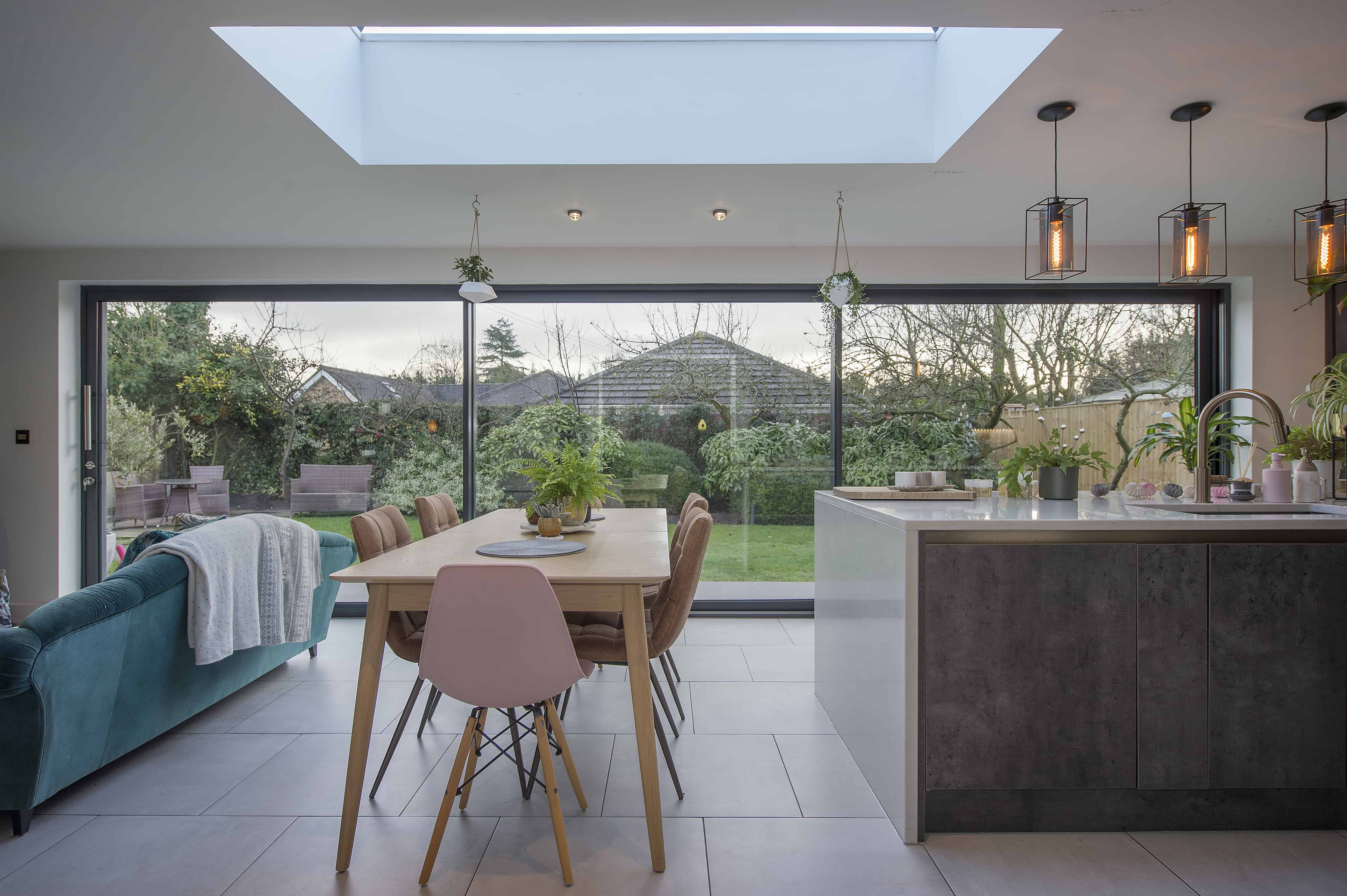 Modern open-plan kitchen and dining area with a skylight, large sliding glass doors overlooking a lush garden, a wooden dining table with pink and wood chairs, and an island with plants and hanging pendant lights.