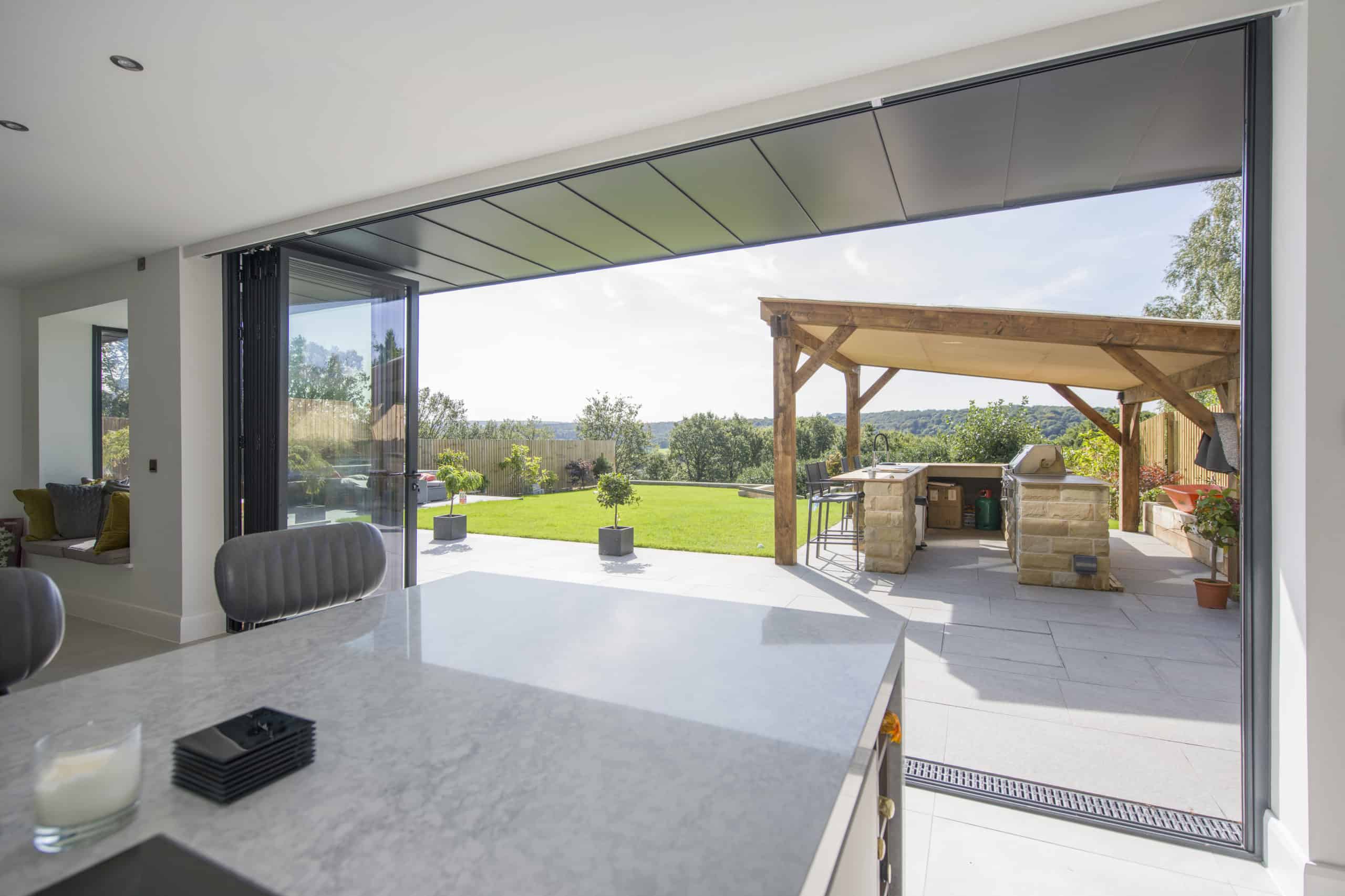 A modern kitchen with a marble countertop opens to a spacious patio and green lawn; outside, there’s a wooden pergola covering an outdoor kitchen and seating area, surrounded by trees and plants.