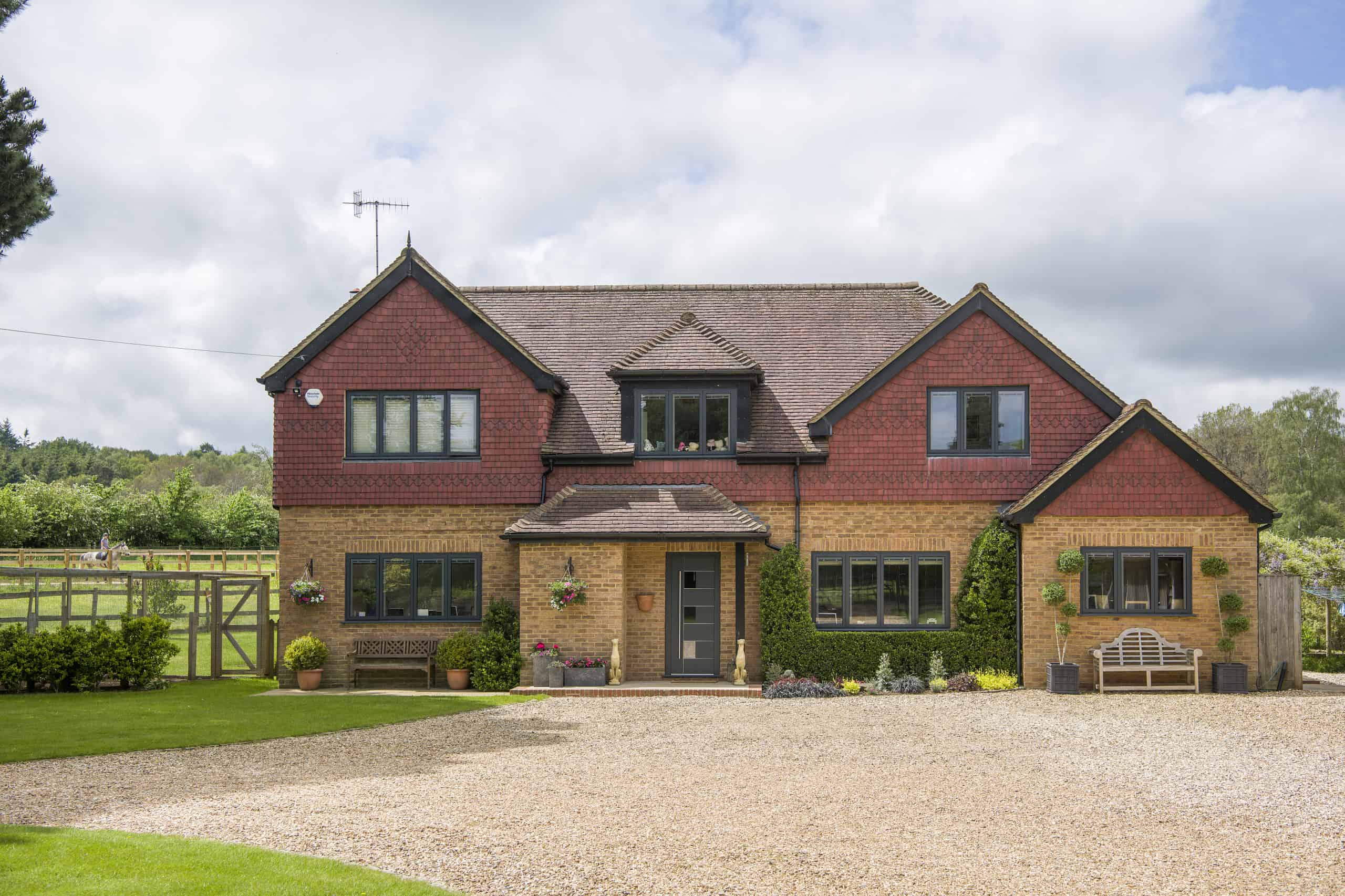 Two-story house with red shingles above yellow brick, a gray front door, large windows, benches outside, and a gravel driveway bordered by gardens—enhanced with bespoke glass solutions for modern appeal.