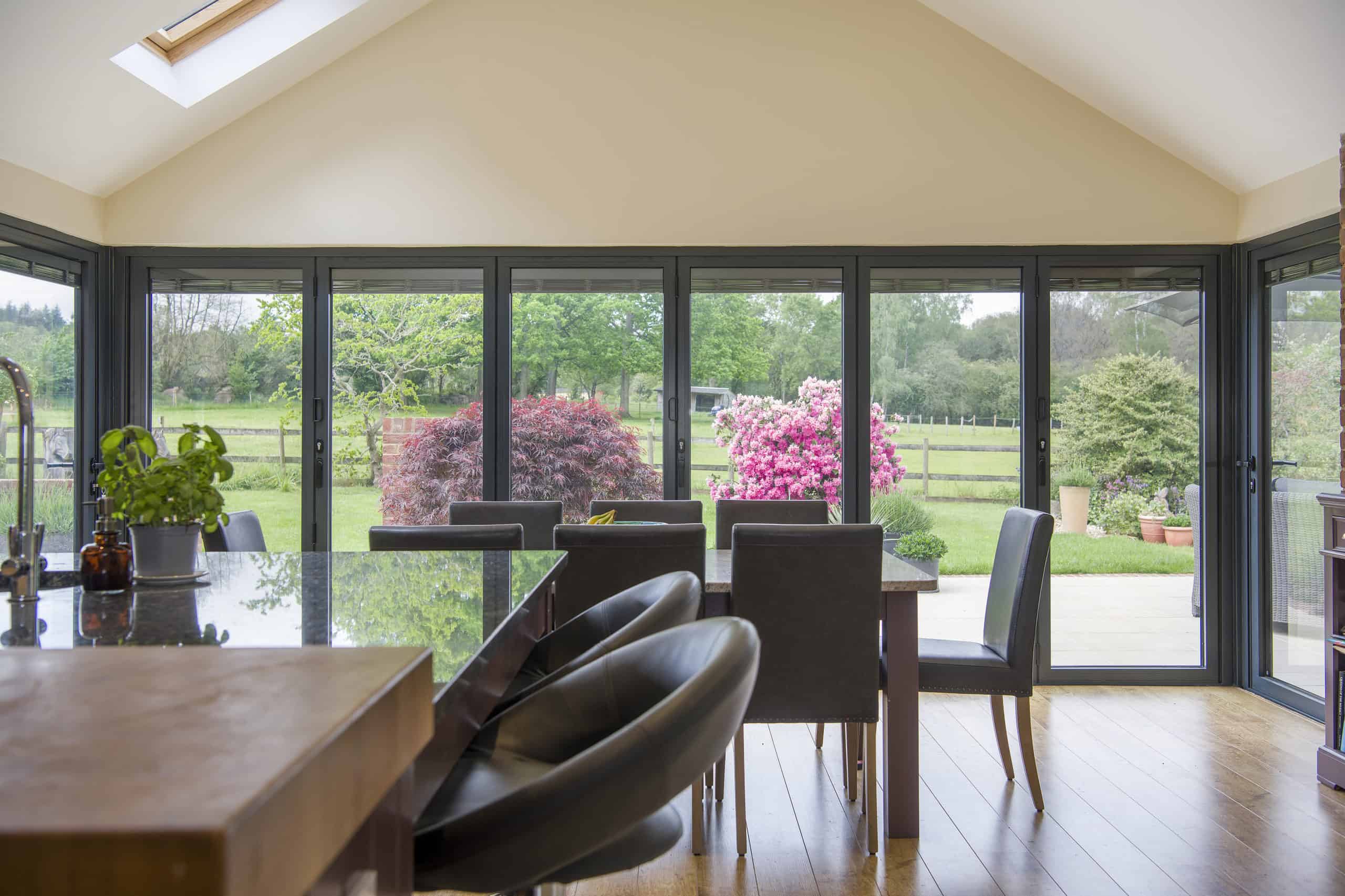 A modern dining area with dark chairs and a glass table faces large folding bespoke glass solutions, opening to a patio and a lush garden with green grass, shrubs, and blooming pink flowers. Natural light fills the space.