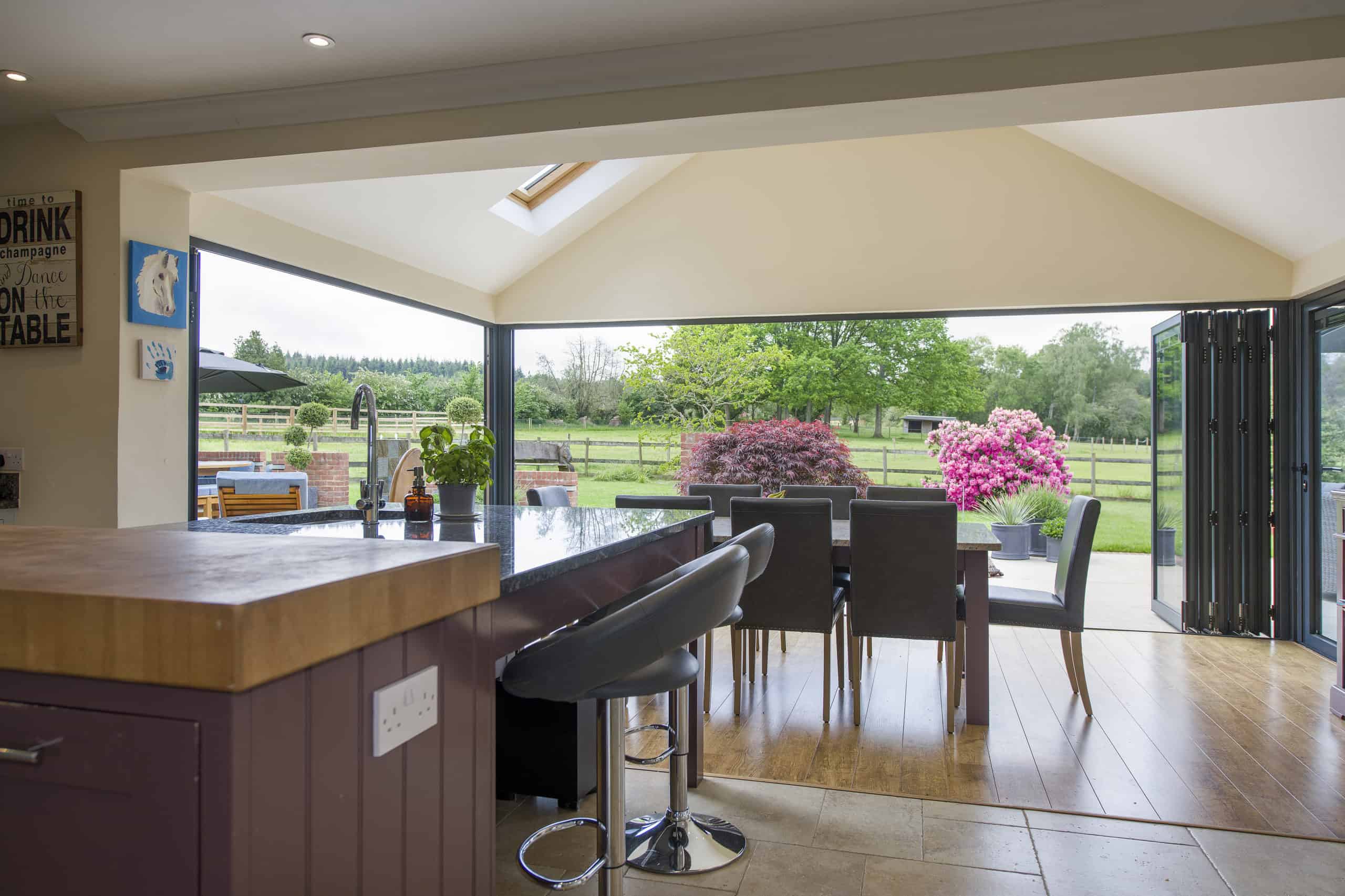 Modern open-plan kitchen and dining area with large bifold doors opened to a patio, revealing a vibrant garden with trees, green grass, and blooming pink bushes. Natural light fills the bright, airy space.