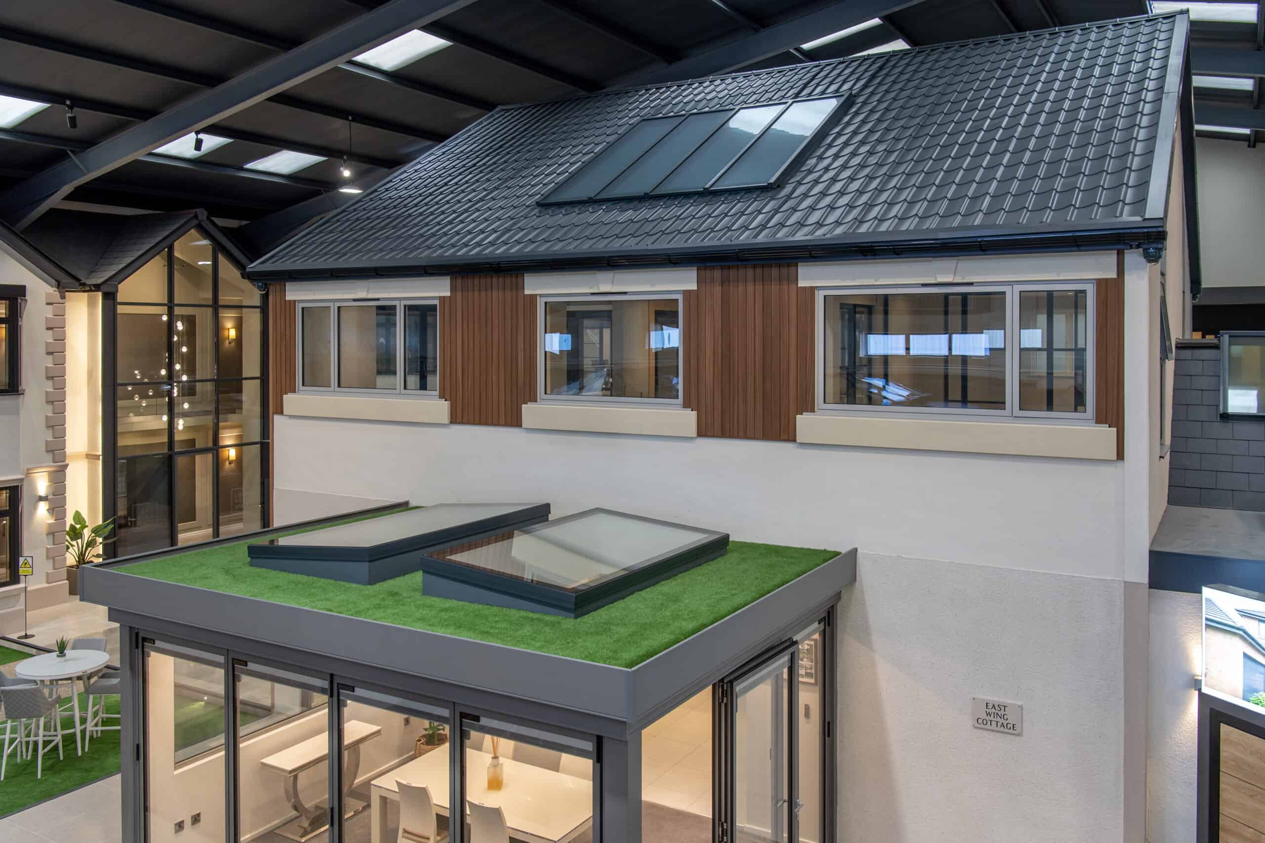 Modern indoor showroom featuring a two-story house model with a gray tiled roof, large skylights, wood and cream exterior, and bespoke glass solutions like bifold doors and glass roofing over a green rooftop patio area.