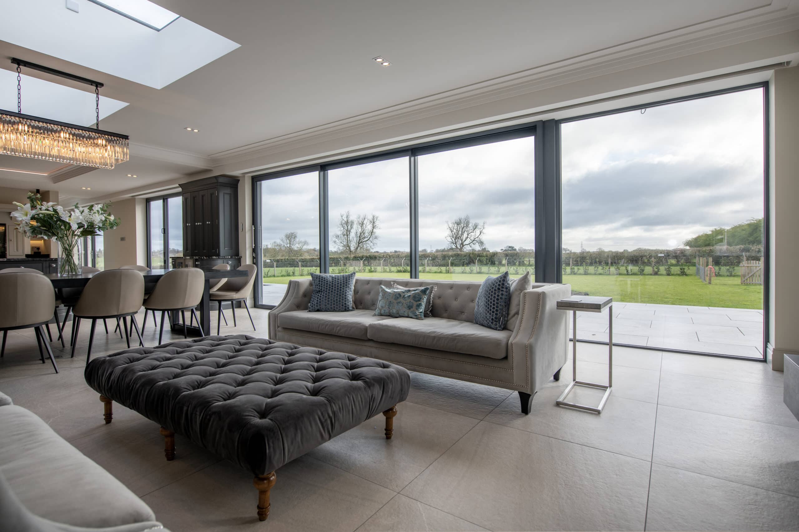 Modern open-plan living area with large bifold doors, grey sofas, a tufted ottoman, dining table with chairs, and a view of a green lawn and trees outside under a cloudy sky.