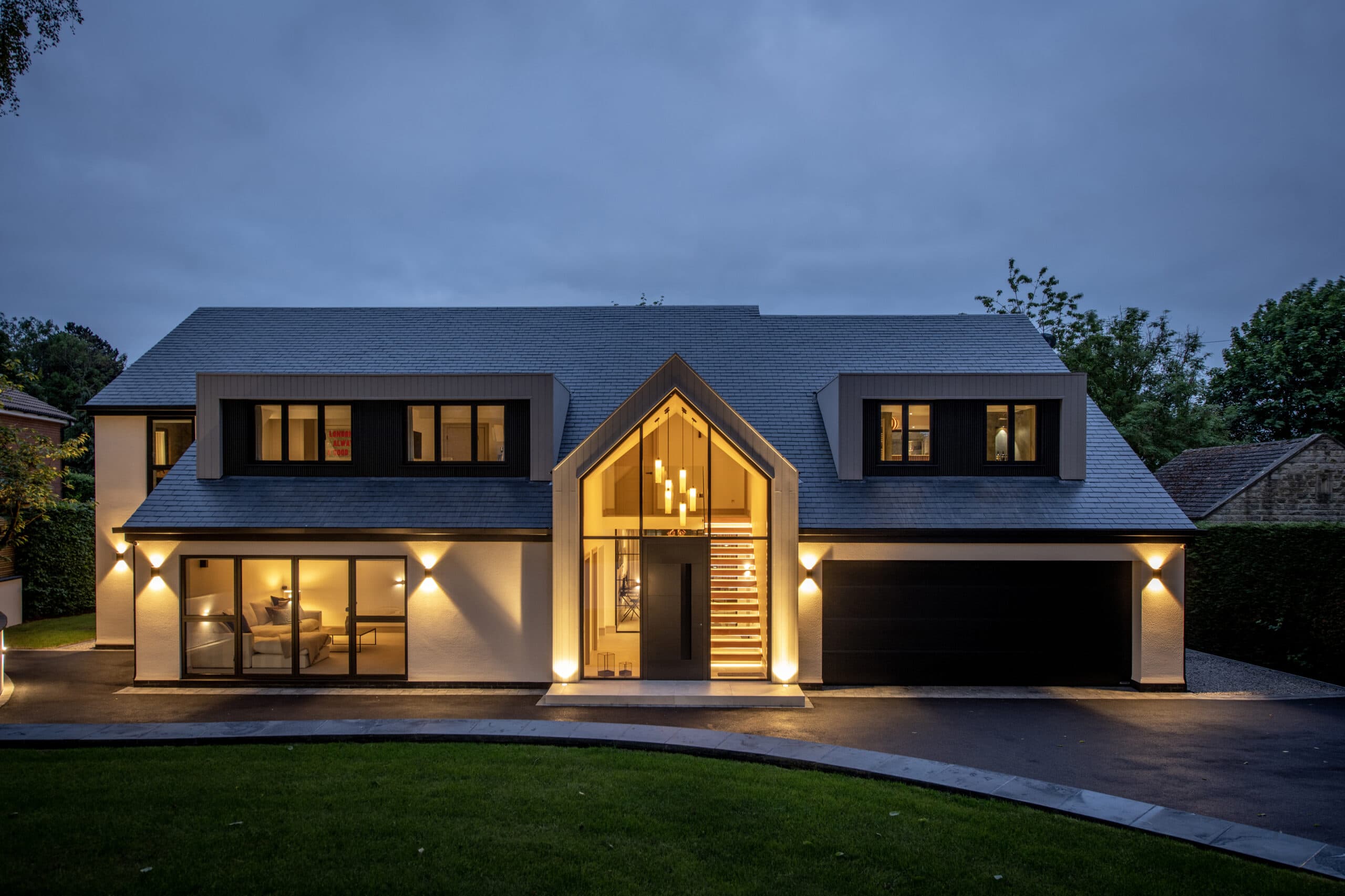 Modern two-story house at dusk with large windows, a striking triangular entryway, bespoke glass solutions, an illuminated staircase, a black garage door, and exterior lights highlighting the driveway and lawn.