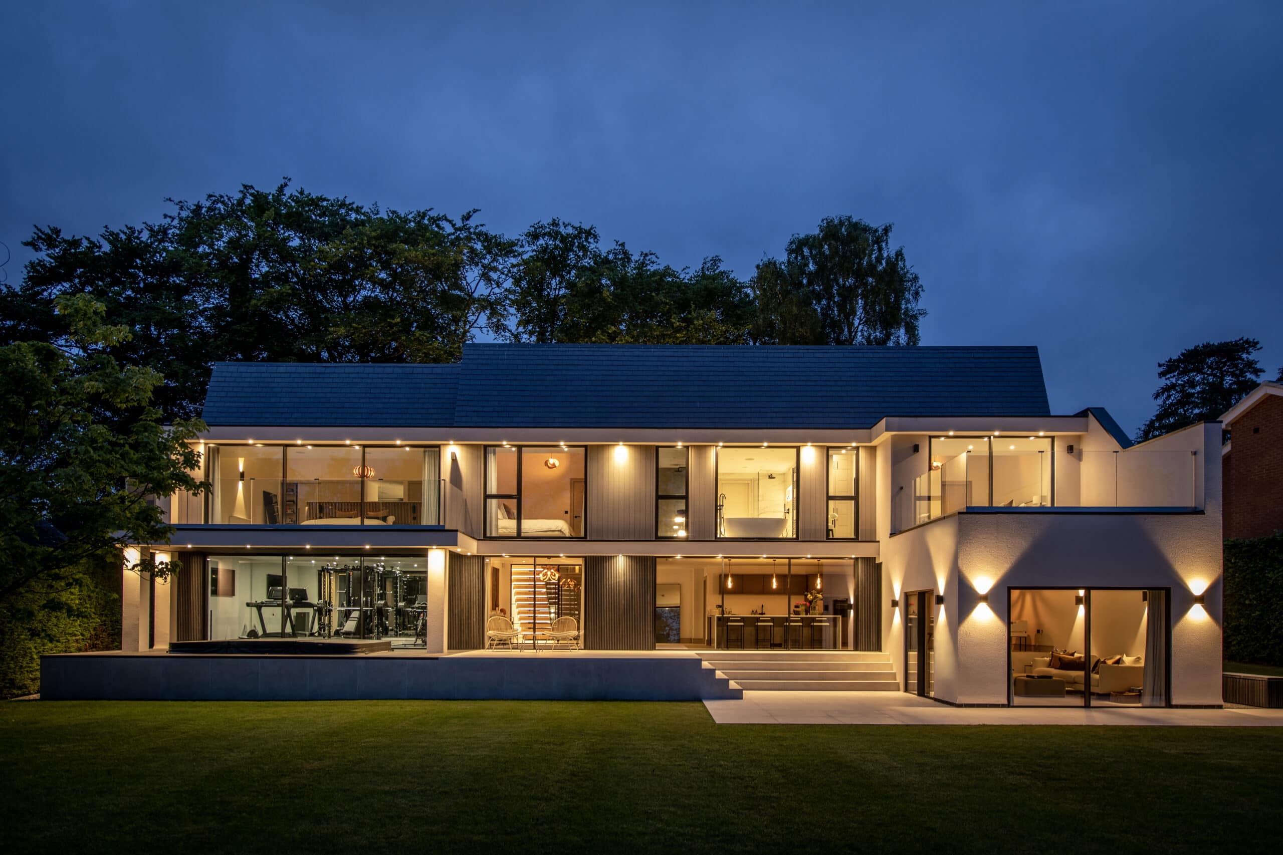 A modern two-story house with large glass windows and sliding doors is illuminated at dusk. The interior lights reveal spacious rooms, including a gym, and the home is surrounded by trees and a well-manicured lawn.