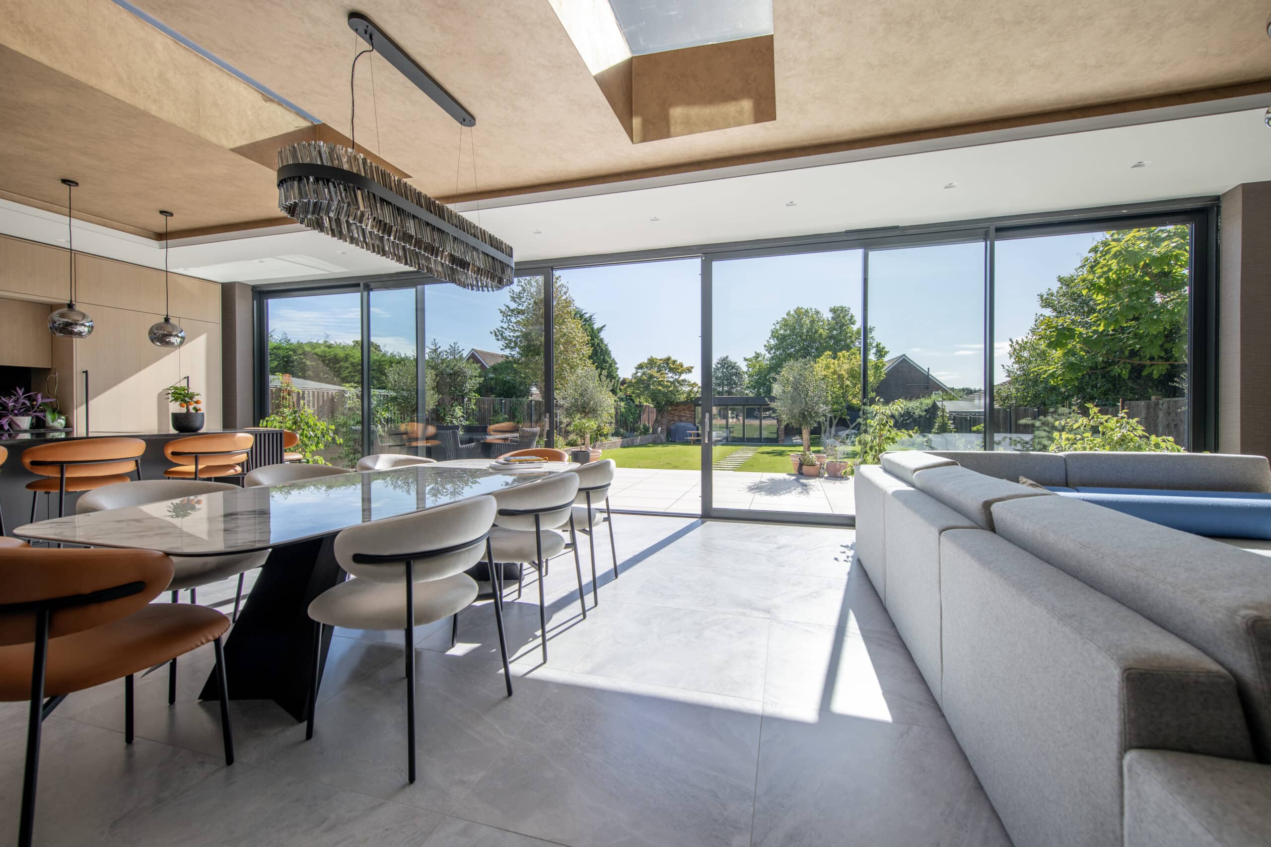 Modern dining and living area with large windows and sliding doors opening to a sunny garden. A marble dining table with stylish chairs sits near a gray sectional sofa, all in a bright, open-plan space with views of greenery outside.