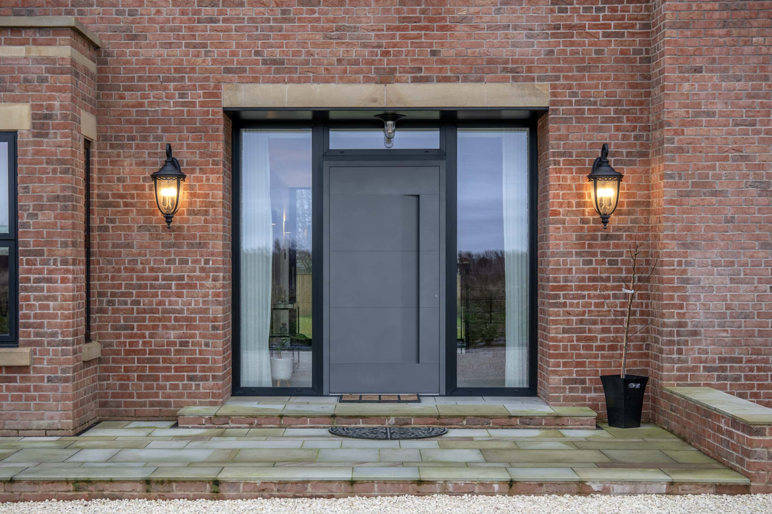 Modern gray front door with vertical bespoke glass solutions on both sides, set in a red brick exterior wall. Two lit lantern-style wall lights flank the entrance, with a stone-tiled doorstep and a doormat.
