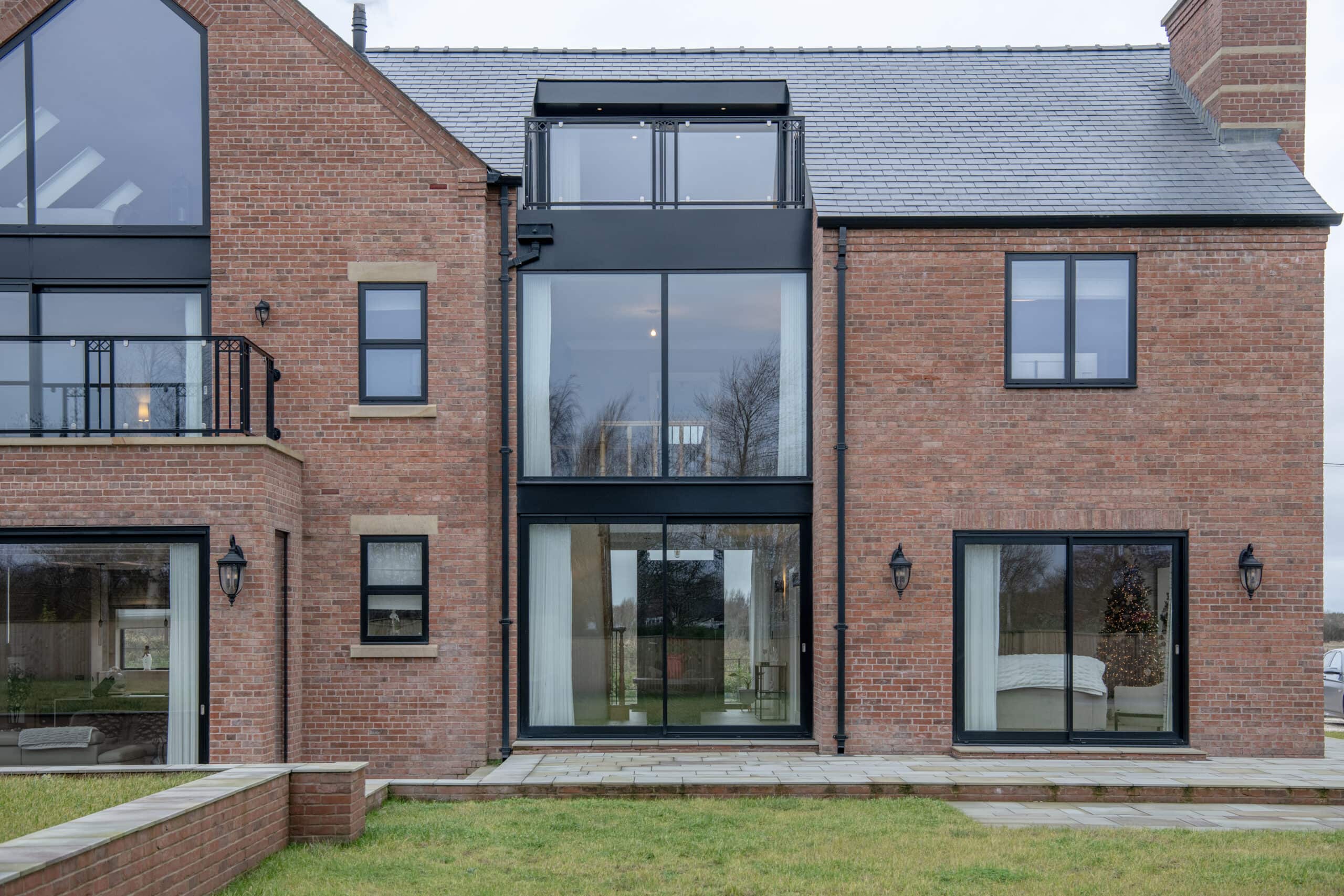 A modern brick house with large glass windows and sleek black frames. Sliding doors open onto a patio by the entrance. A well-kept lawn stretches out front, while a Christmas tree glows indoors through one window.