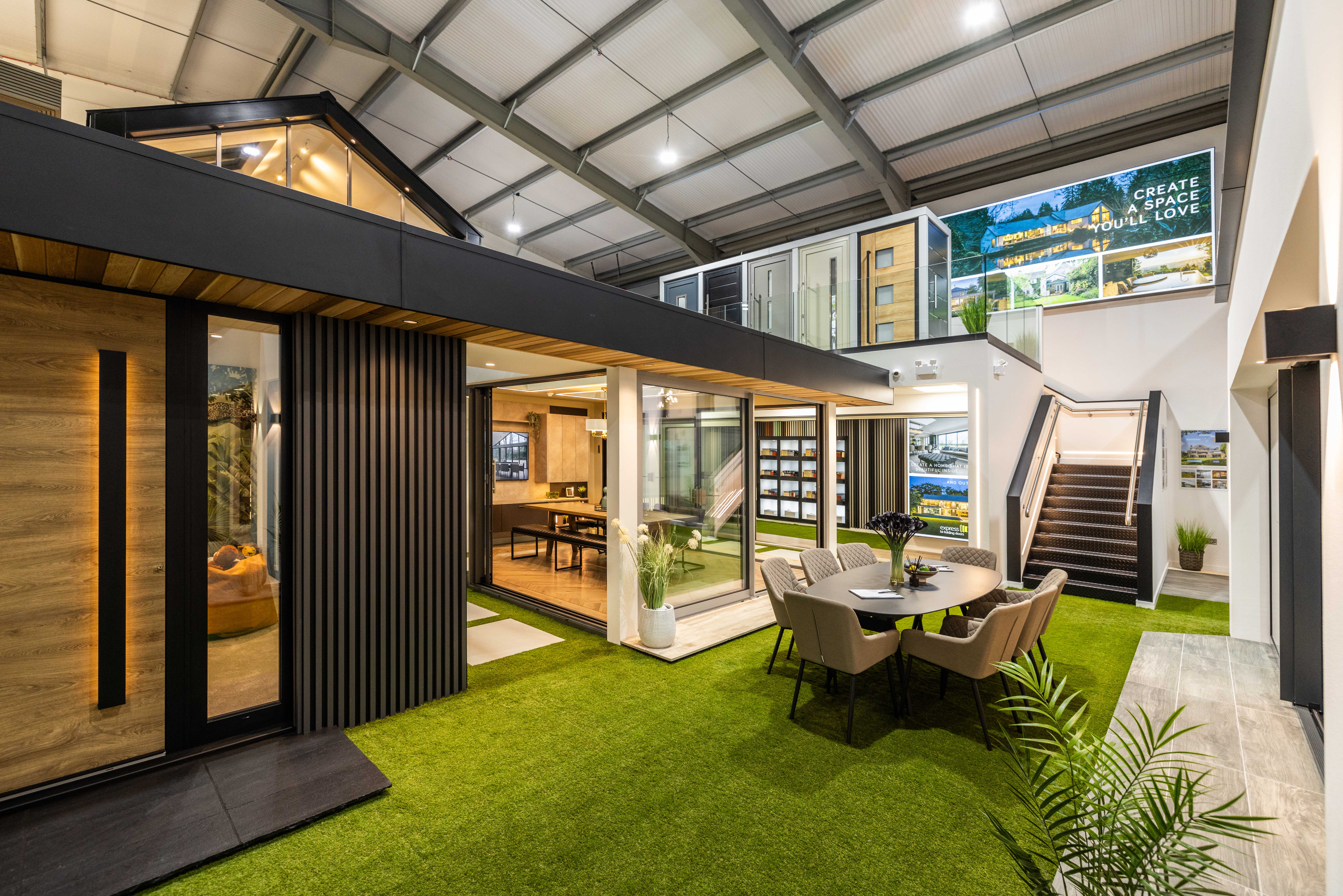 A modern, open-concept indoor showroom features modular home displays, large windows with bespoke glass solutions, a dining table set, greenery, and stairs leading to an upper level. The space is well-lit with a high, exposed ceiling.