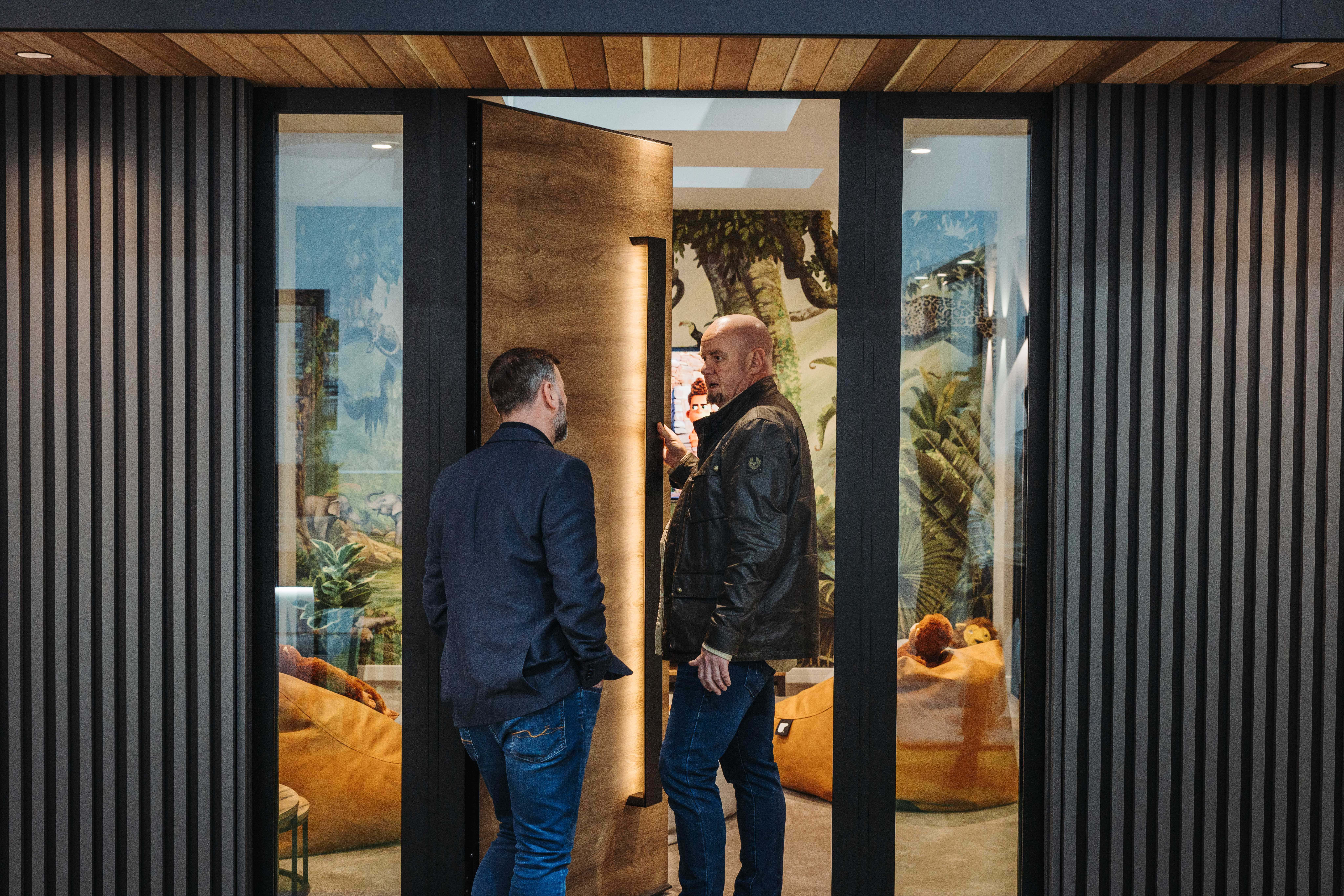 Two men stand at the entrance of a modern building, one opening large bifold front doors while the other talks to him. Through the doorway, cozy seating and lush plants are visible under stylish glass roofing.