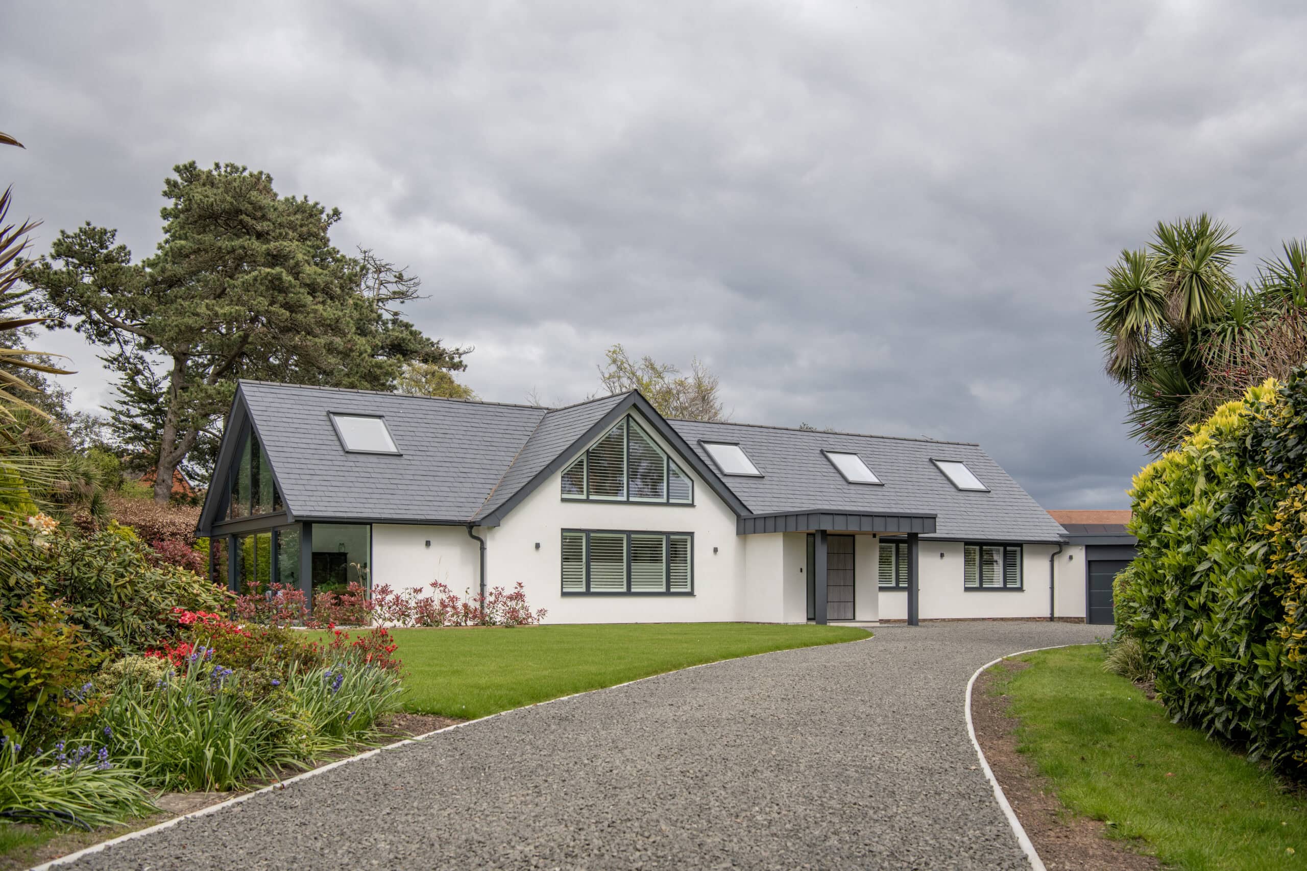 Modern single-story house with large windows, sliding doors, and a gray roof, surrounded by green lawn, colorful flowers, and trees under a cloudy sky with a gravel driveway leading to the front door.