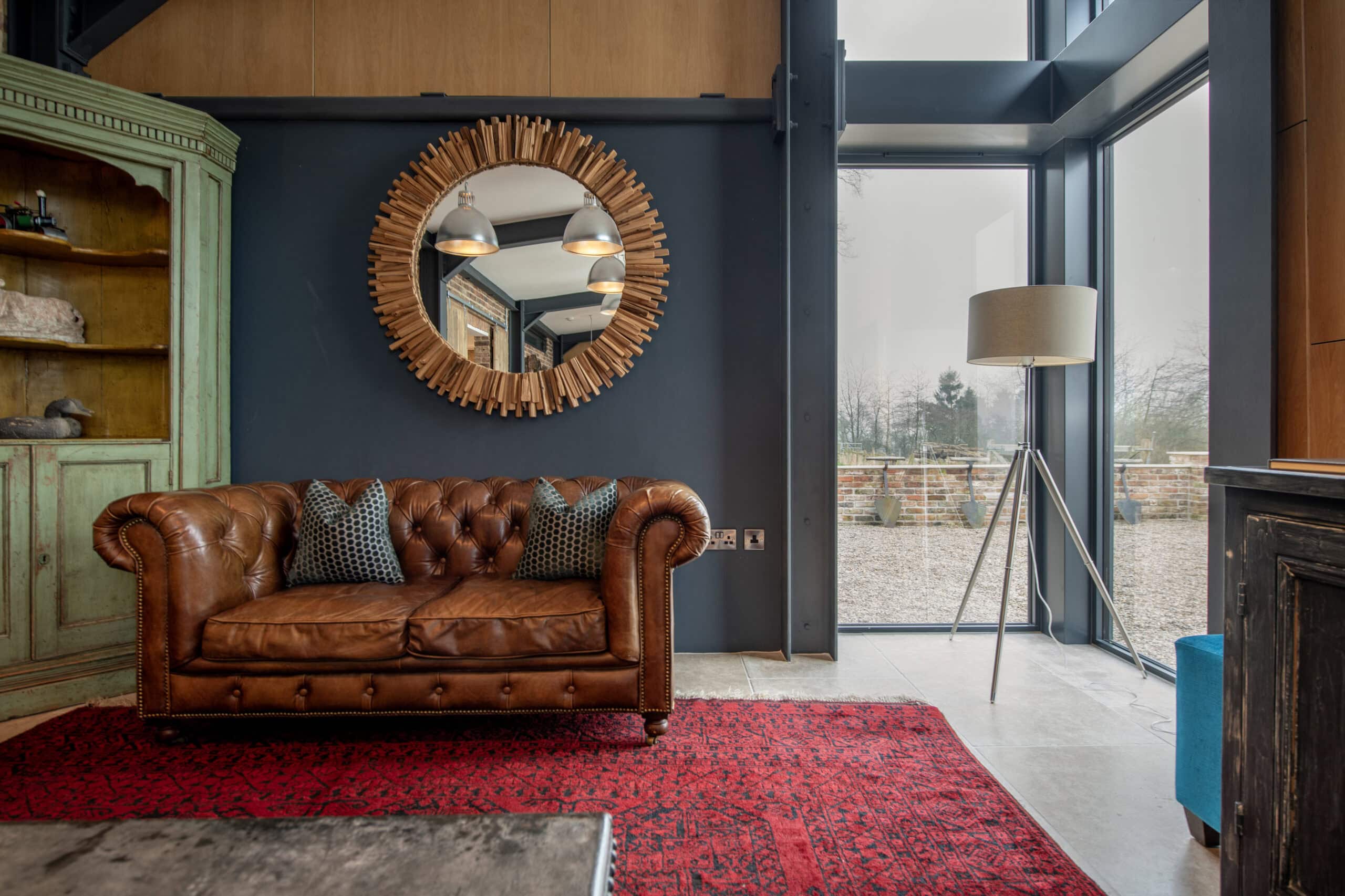 A cozy living room with a brown leather sofa, two patterned pillows, a red rug, a round wooden-framed mirror on a dark wall, a tall floor lamp, and large windows with sliding doors letting in natural light.