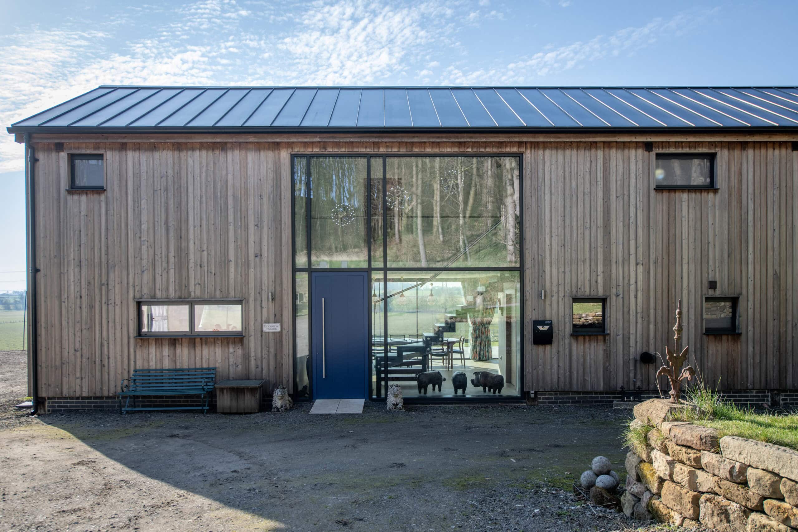 A modern wooden building with large glass windows and a blue door, featuring bespoke glass solutions throughout. A bench and mailbox sit outside, while the upper floors reflect trees and sky. Stones and a small plant are in the foreground.