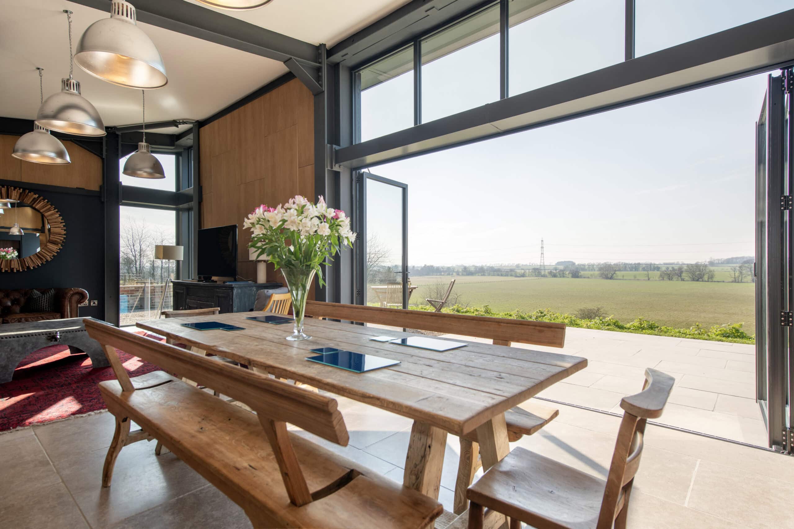 A bright, modern dining area with wooden table and benches, a vase of flowers, and large sliding doors offering a scenic view of a green countryside landscape under a clear sky.