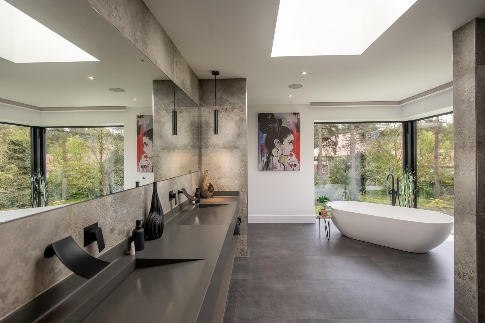 Modern bathroom with large mirrors, double sinks, a freestanding bathtub near tall windows, artwork on the wall, grey floors, glass roofing for extra natural light, and views of surrounding greenery.