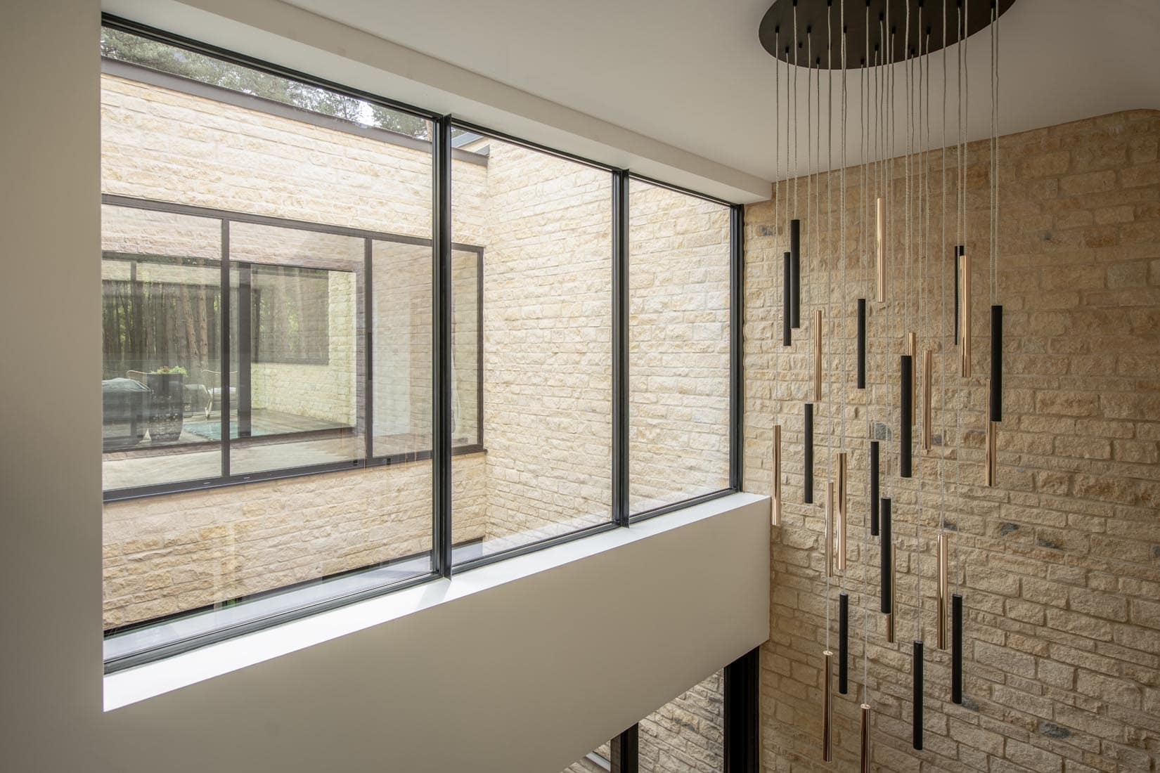 Modern interior with large floor-to-ceiling windows, beige stone walls, and a vertical hanging light fixture with multiple slim rods. Bespoke glass solutions frame the view onto a partially enclosed courtyard.