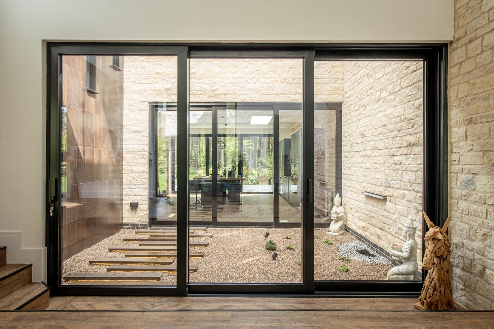 Large sliding doors reveal a modern indoor courtyard with gravel, stepping stones, minimal plants, and two white statues against beige stone walls; natural light fills the space.