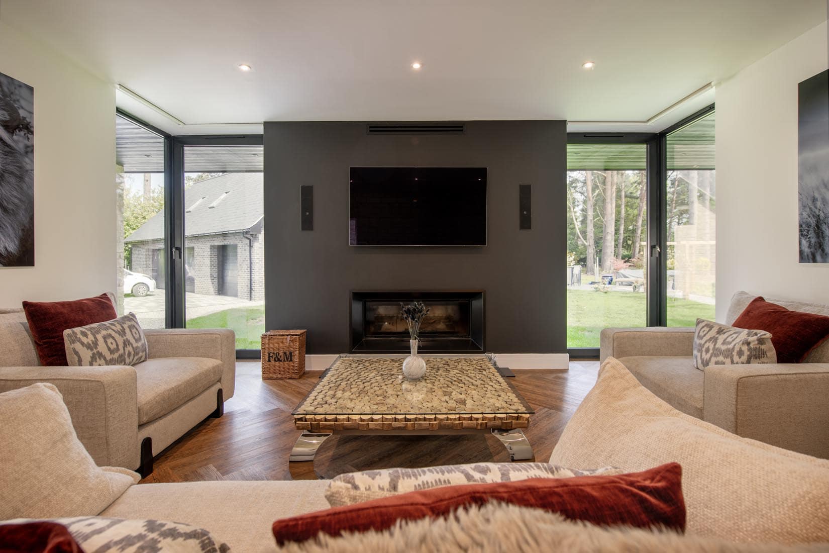 Modern living room with beige sofas, red and patterned cushions, a square glass coffee table, wall-mounted TV, built-in fireplace, and large windows with sliding doors showing greenery outside.