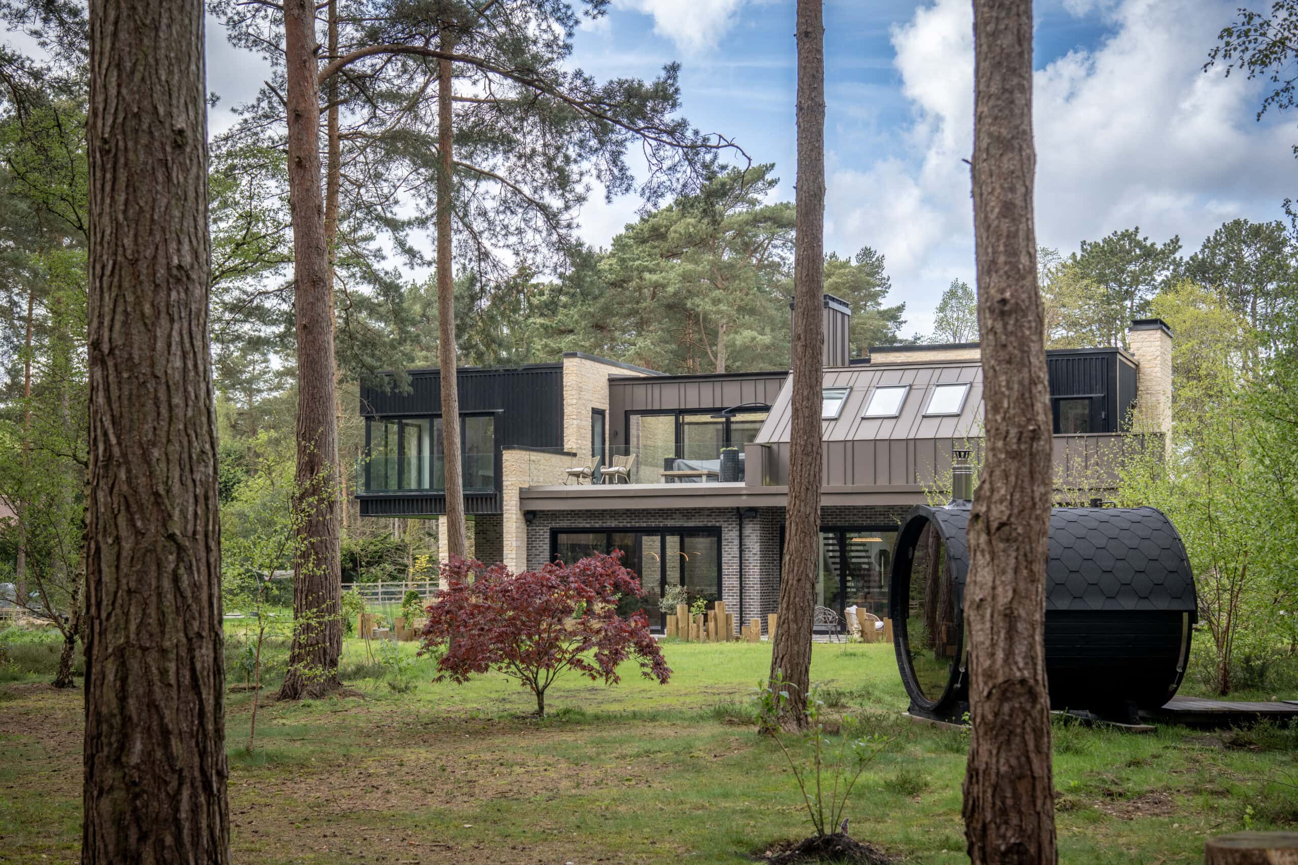 Modern two-story house with large windows, bifold doors, and brick exterior surrounded by tall pine trees and greenery; a small red-leafed tree and a black, barrel-shaped outdoor structure are visible in the yard.