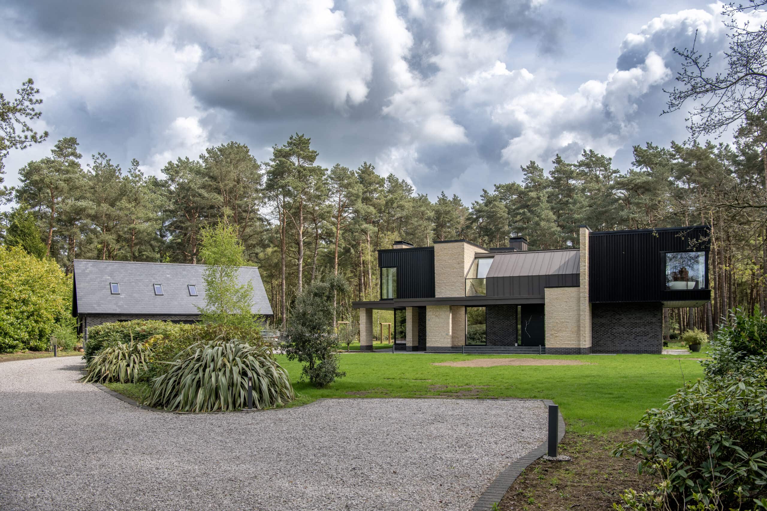 Modern two-story house with large windows, sliding doors, and a black and beige exterior, situated next to a smaller building with a slate roof, surrounded by trees and greenery under a cloudy sky. Gravel driveway in the foreground.