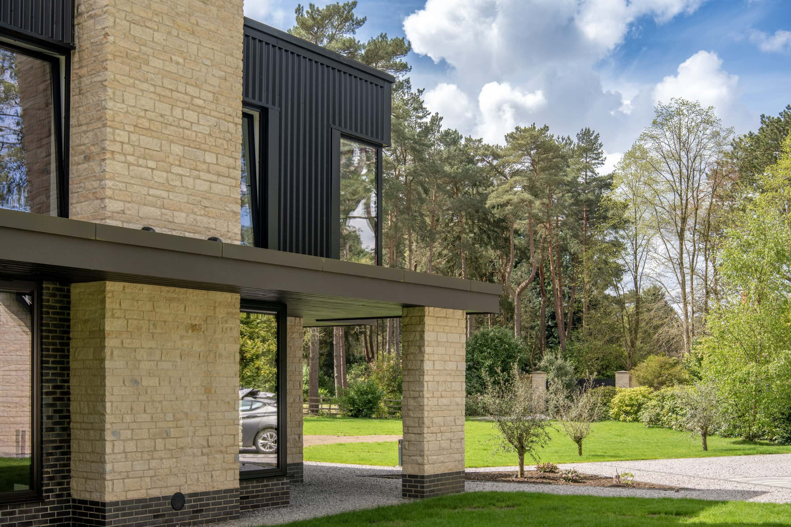 Modern brick house with large windows and sleek bifold doors, featuring a black roof, set in a lush green yard with trees and a car visible in the background under a partly cloudy sky.